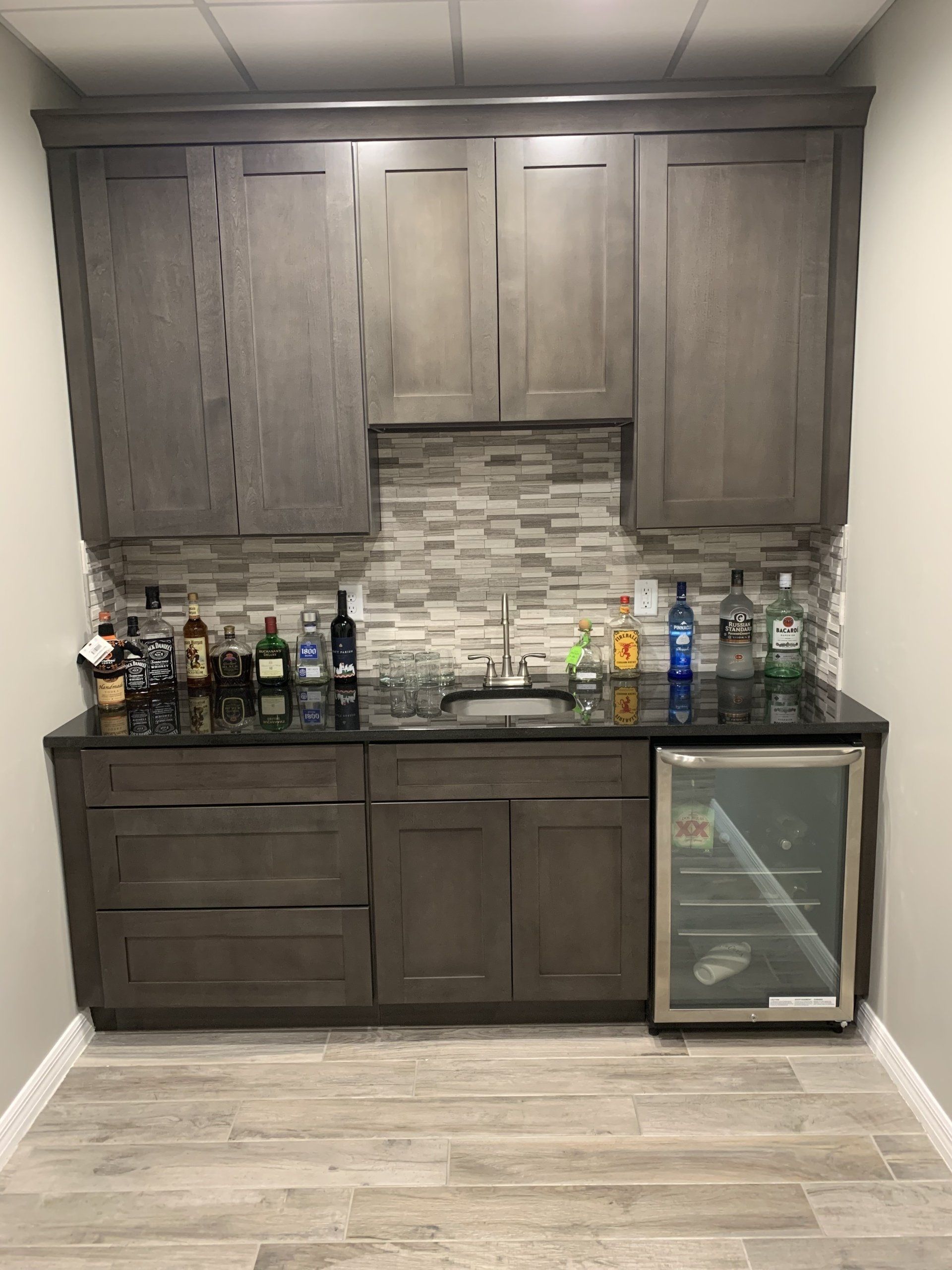 Gray wet bar with upper cabinets, sink, and wine fridge. Dark countertop, tile backsplash, and liquor bottles.