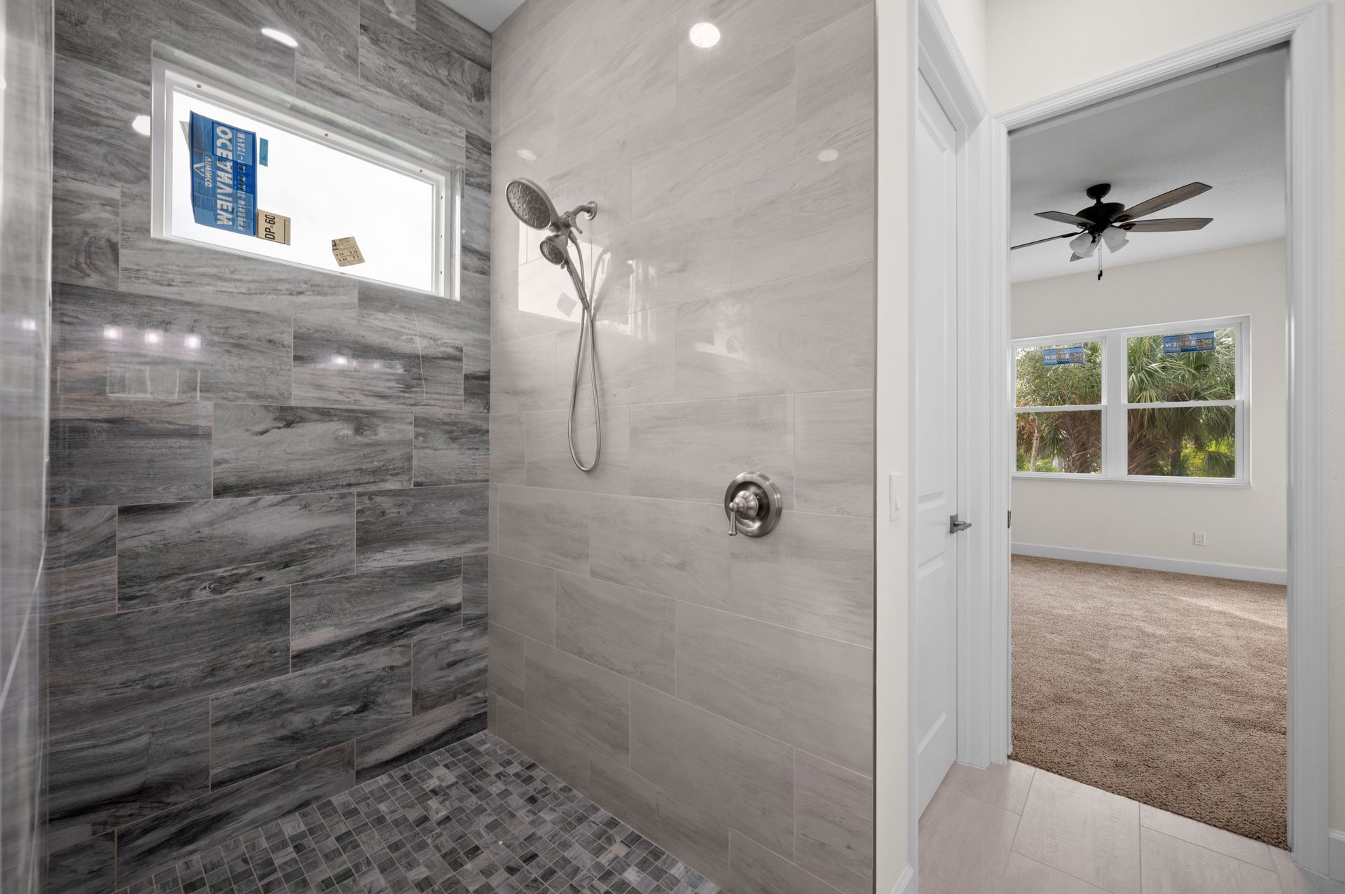 Modern shower with gray tile, pebble floor, and doorway to a bedroom.