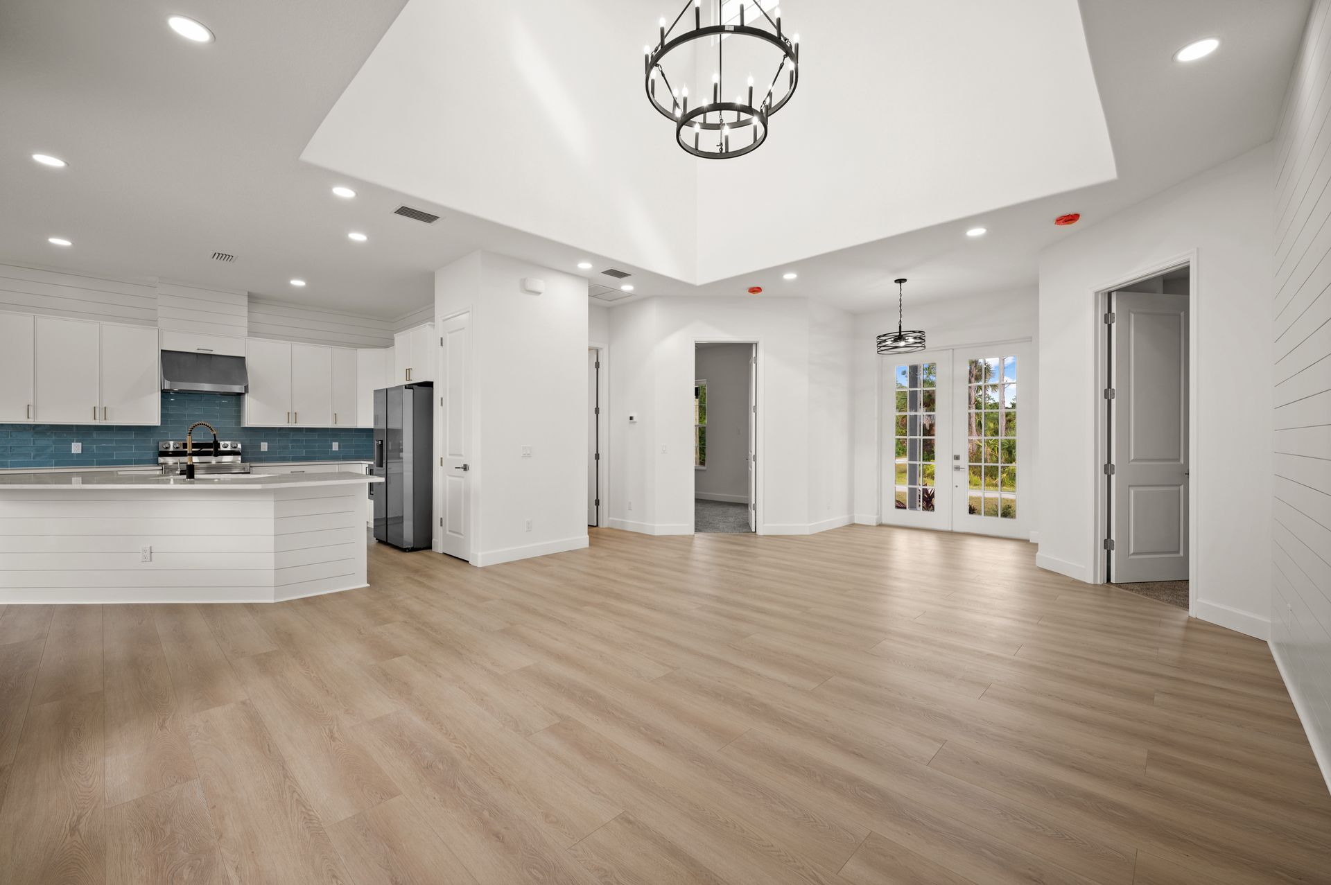 Spacious white interior with a kitchen, French doors, and a chandelier. Wooden floors and lots of natural light.