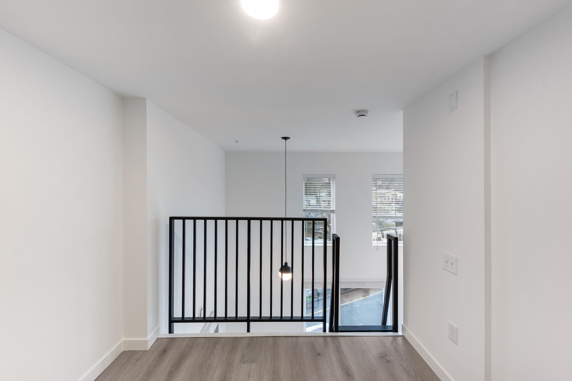 Hallway with white walls, gray floor, black railing, and open space leading to windows.