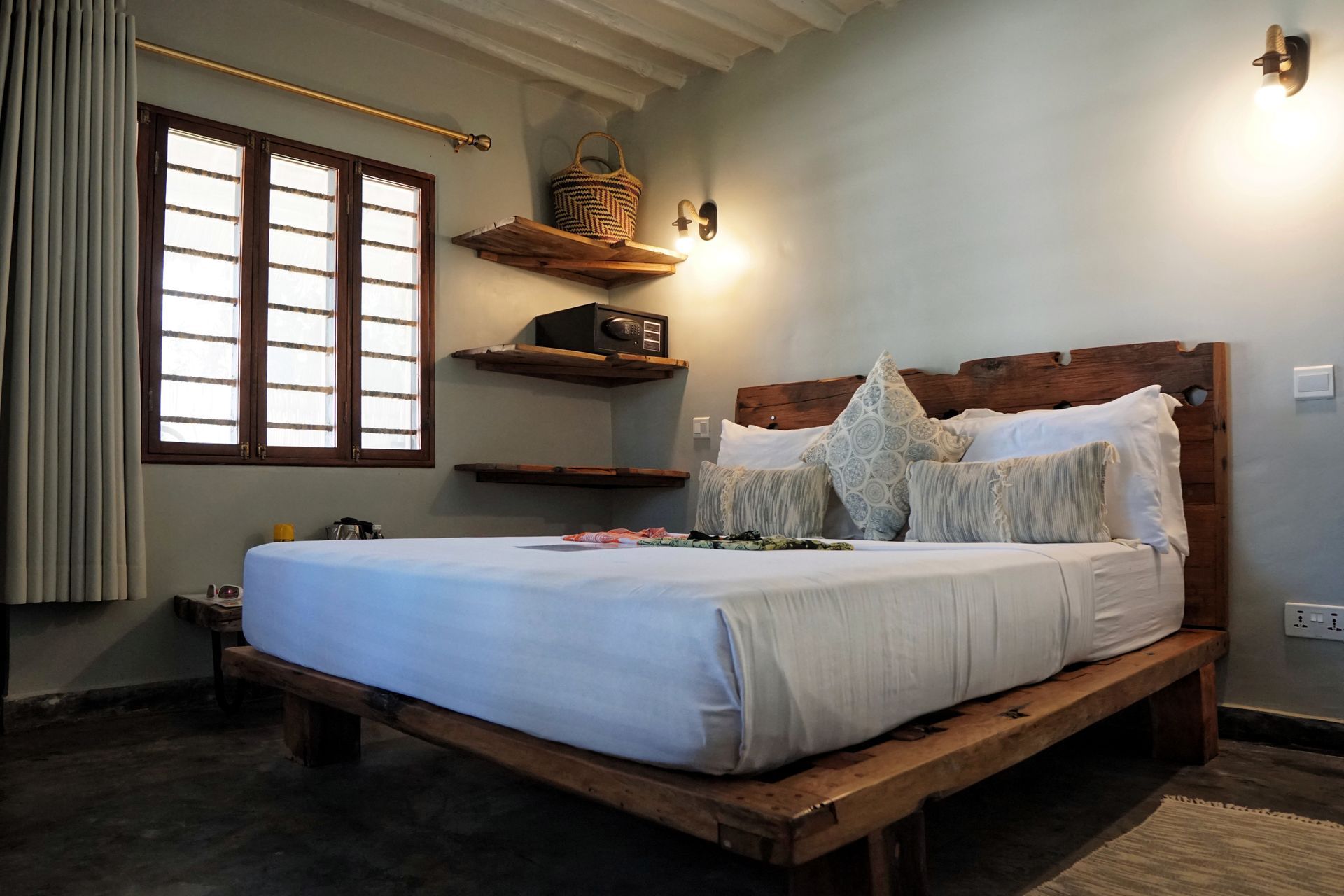 Cozy bedroom with a wooden bed, white bedding, wall shelves, and a window with dark shutters