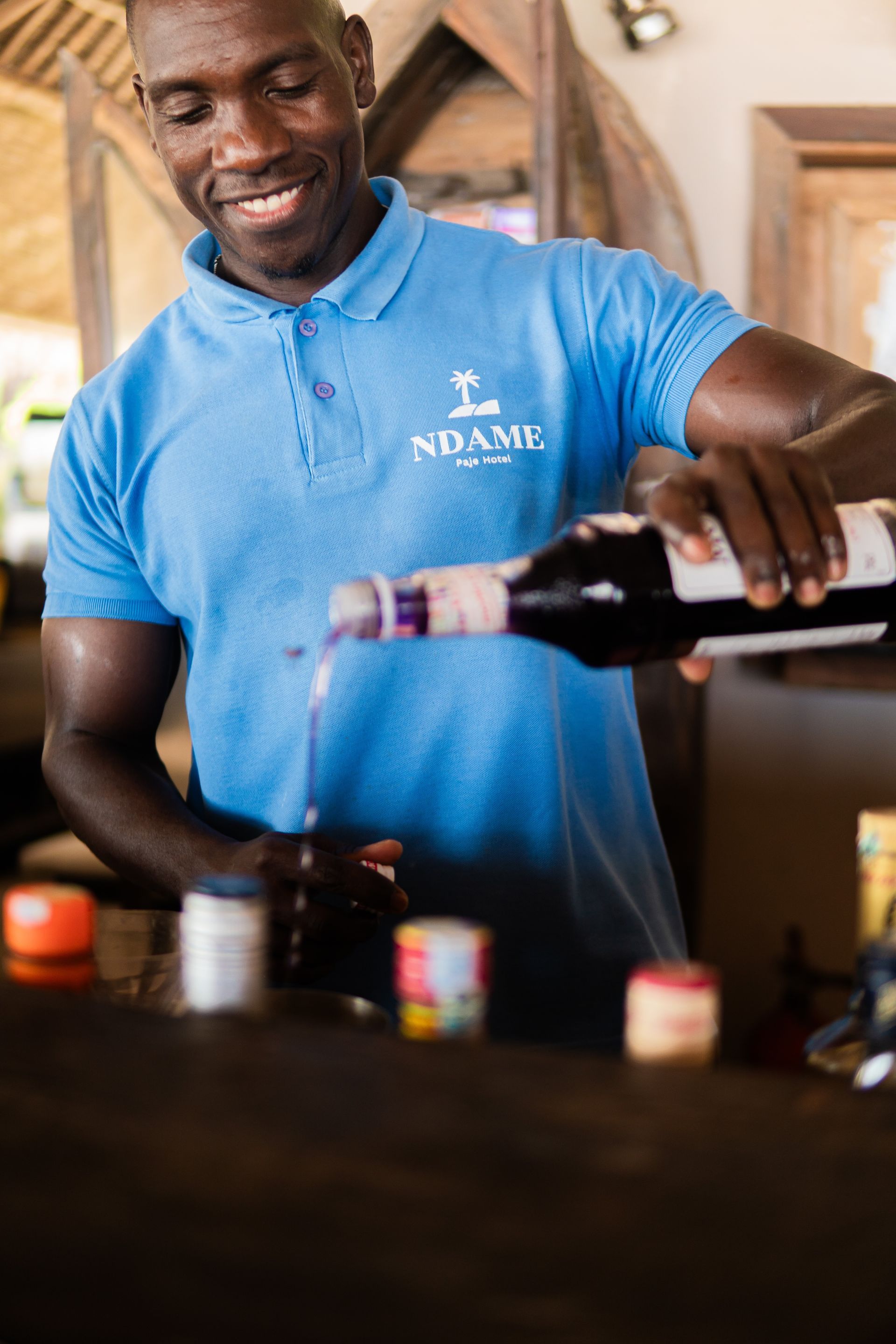 Man pouring wine into a glass beside several bottles on a table, smiling in a bright room