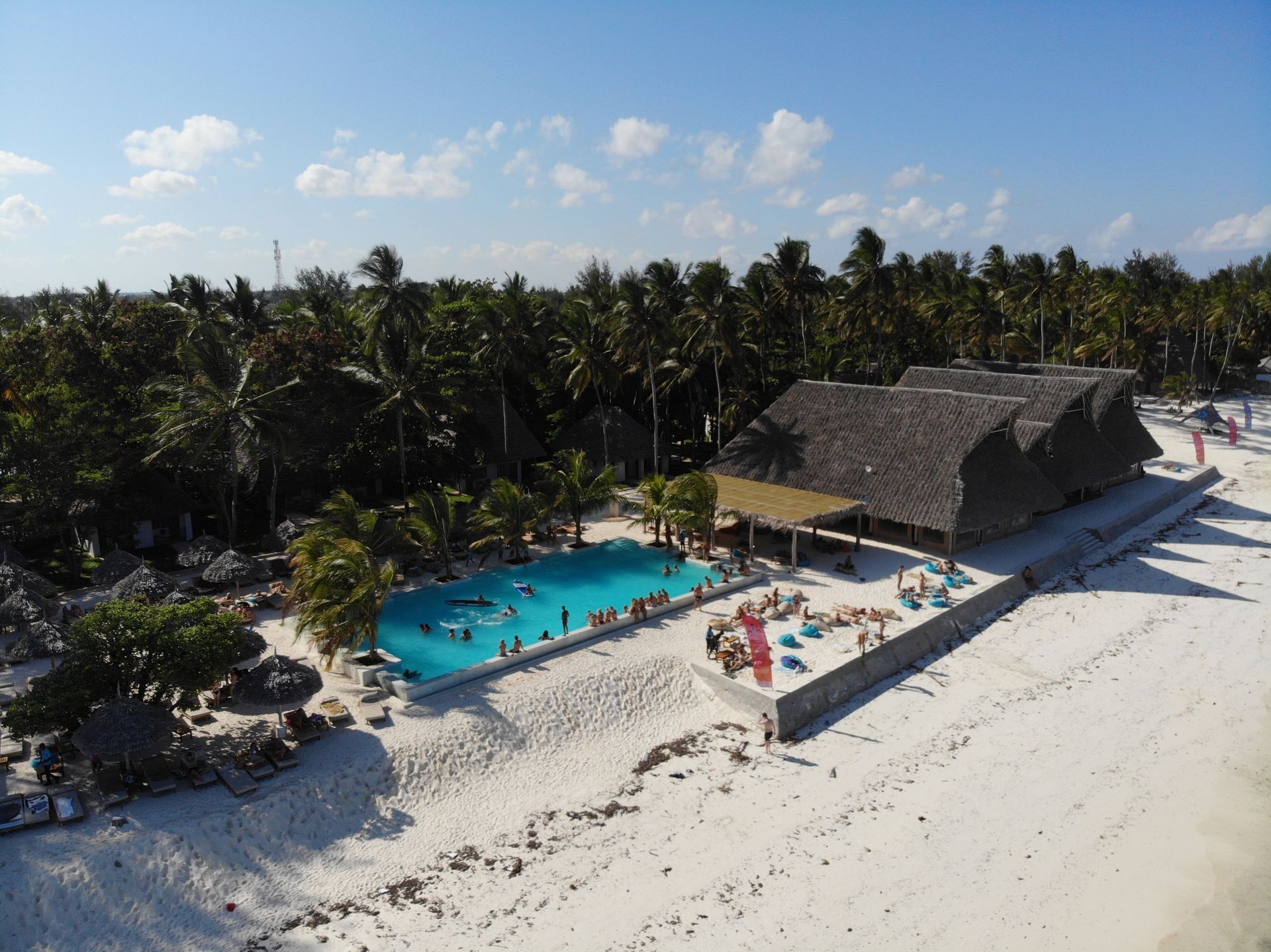 Aerial view of a beach resort with a blue pool, thatched-roof building, palm trees, and white sand.