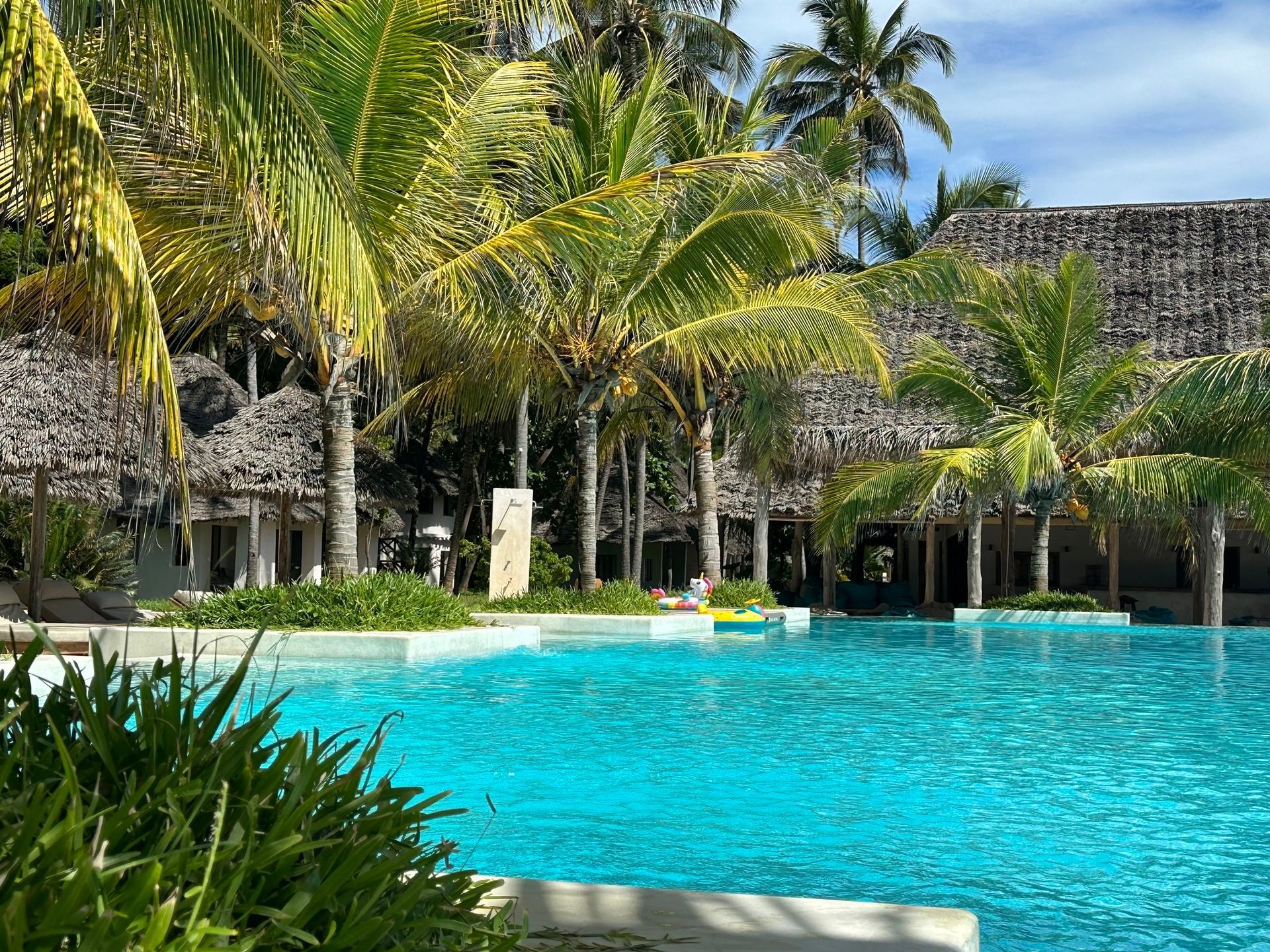 Tropical resort pool with bright turquoise water, palm trees, and thatched-roof buildings under a sunny sky