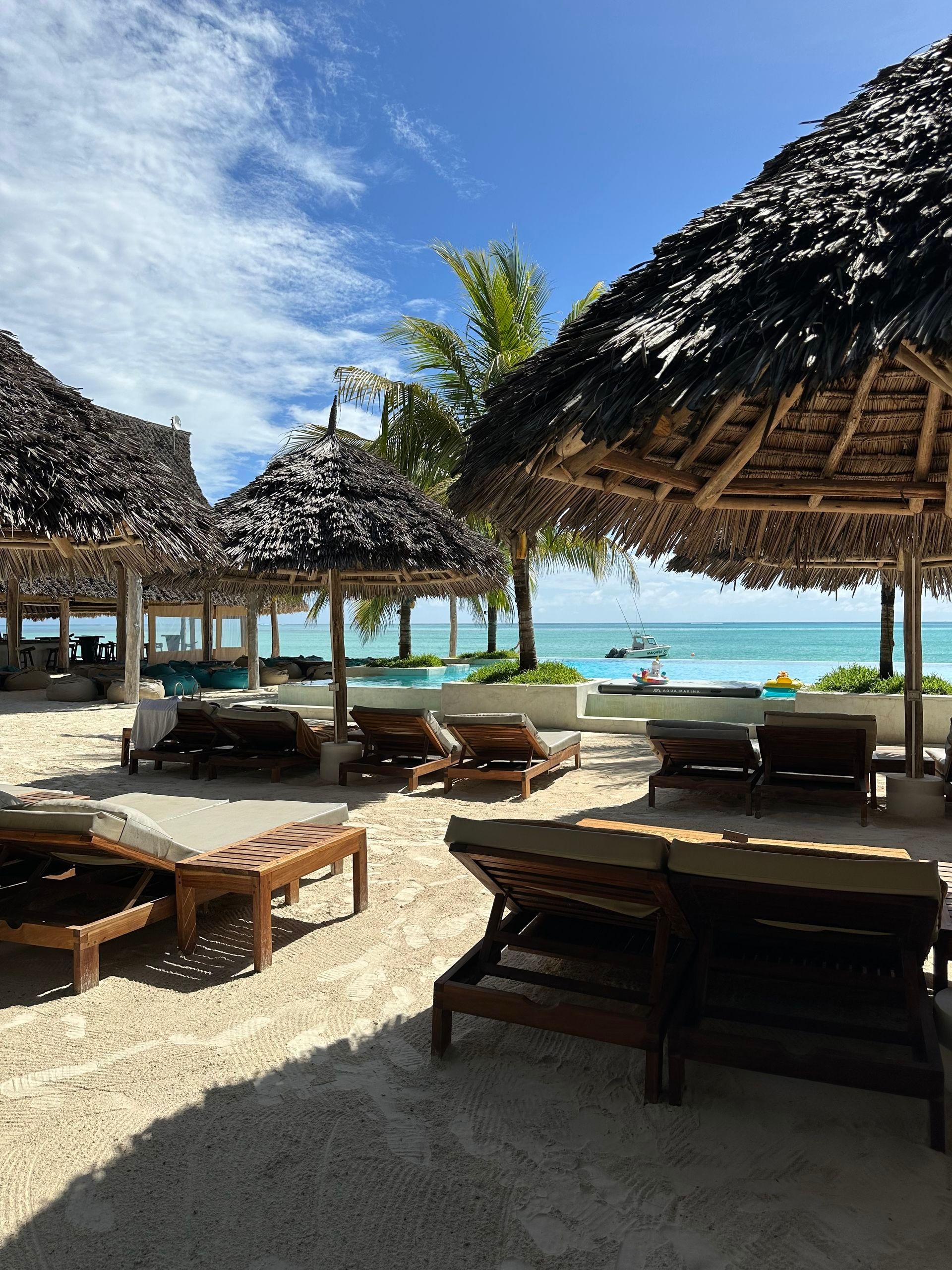 Beach lounge area with thatched-roof cabanas, wooden tables, and ocean view under a blue sky