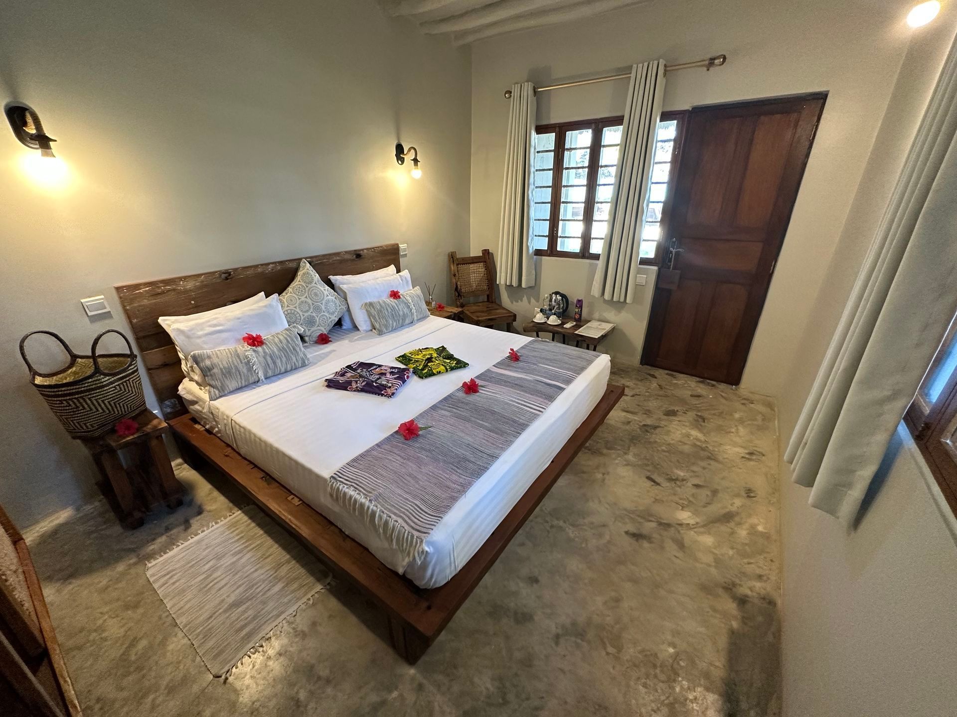 Cozy bedroom with a wooden bed, white linens, wall sconces, window, and wooden door on a tiled floor