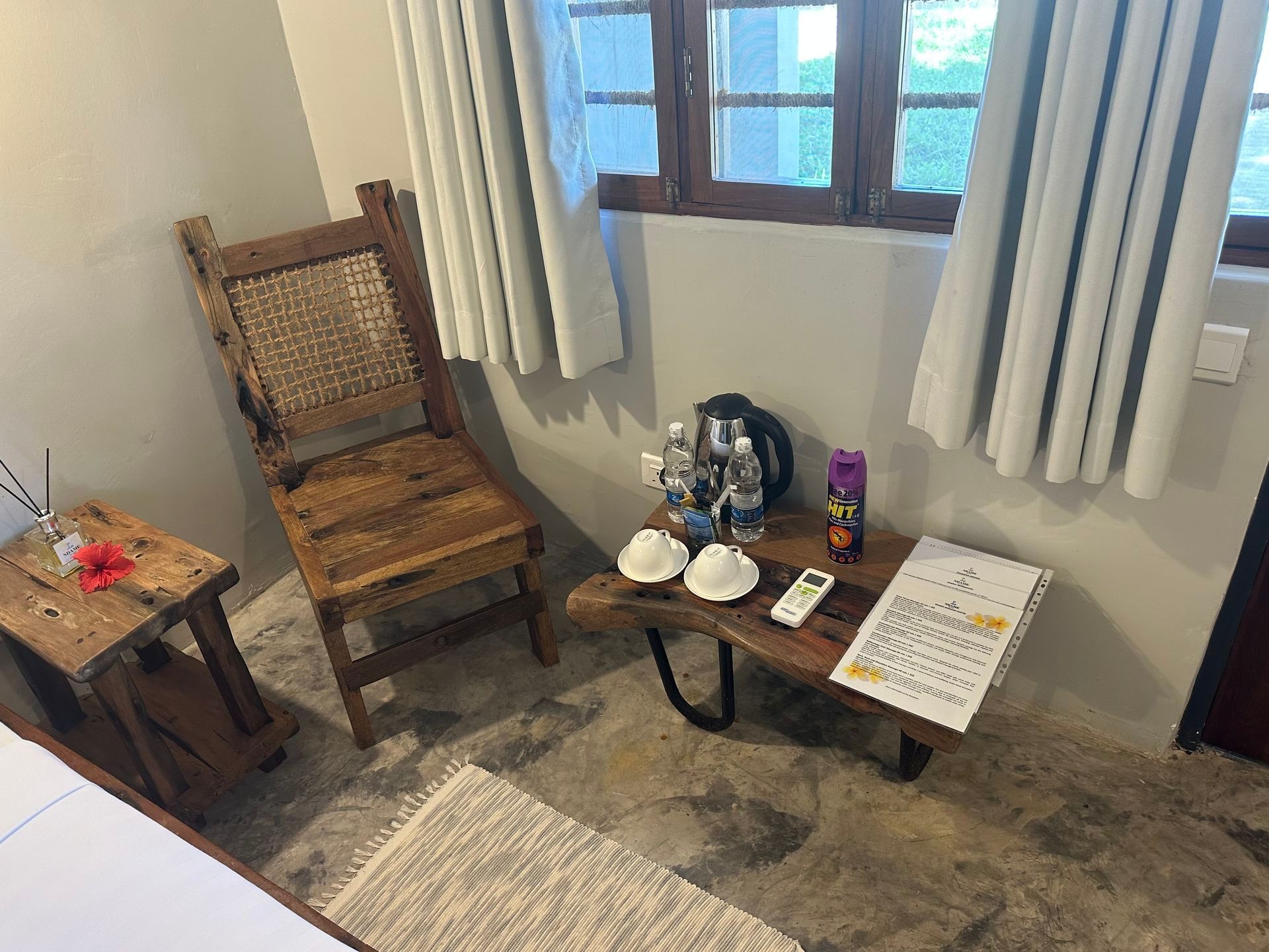 Small room corner with wooden chair, low table, window with curtains, and rug on stone floor.