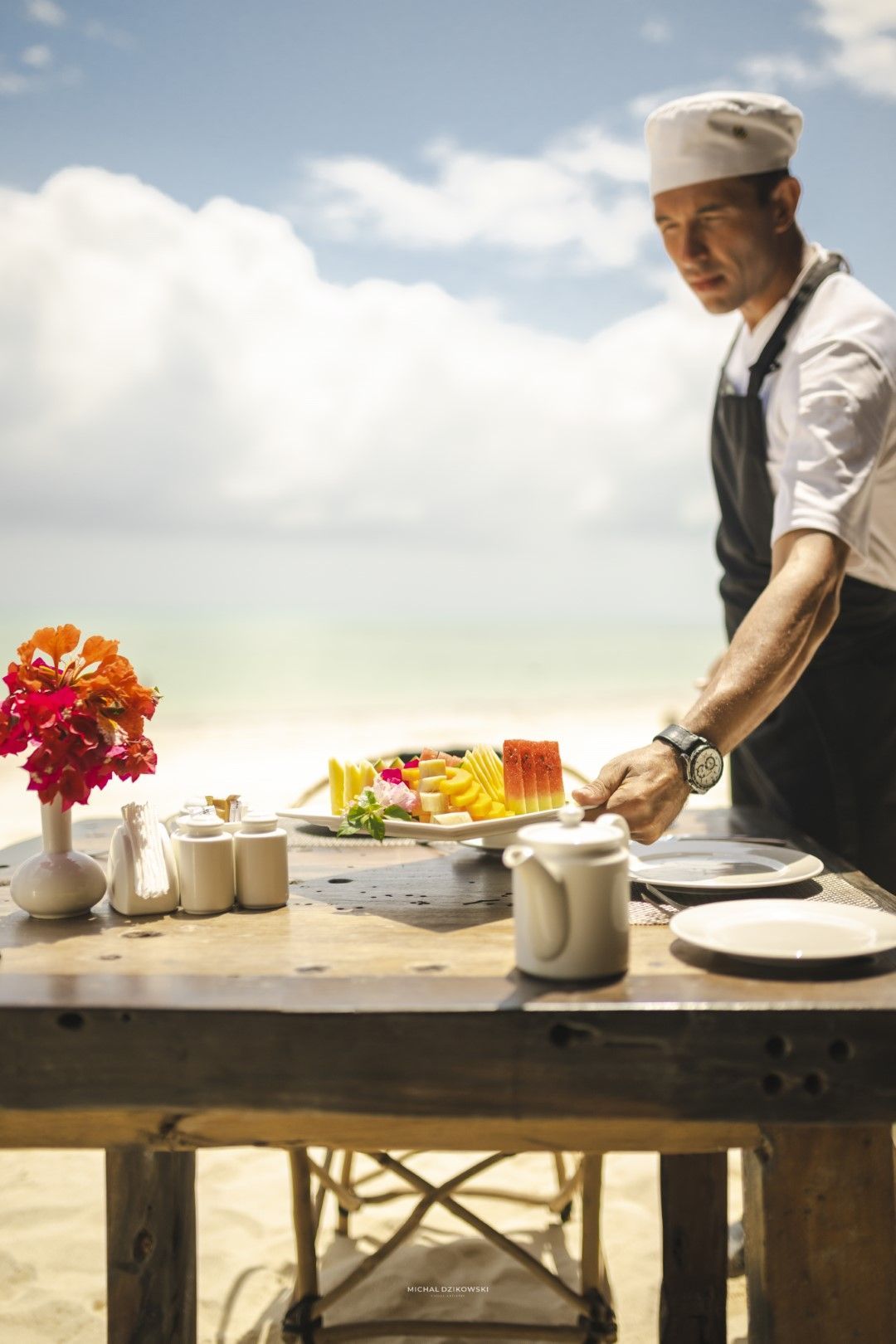 Chef arranging a buffet table outdoors with flowers, drinks, and fruit under a cloudy sky