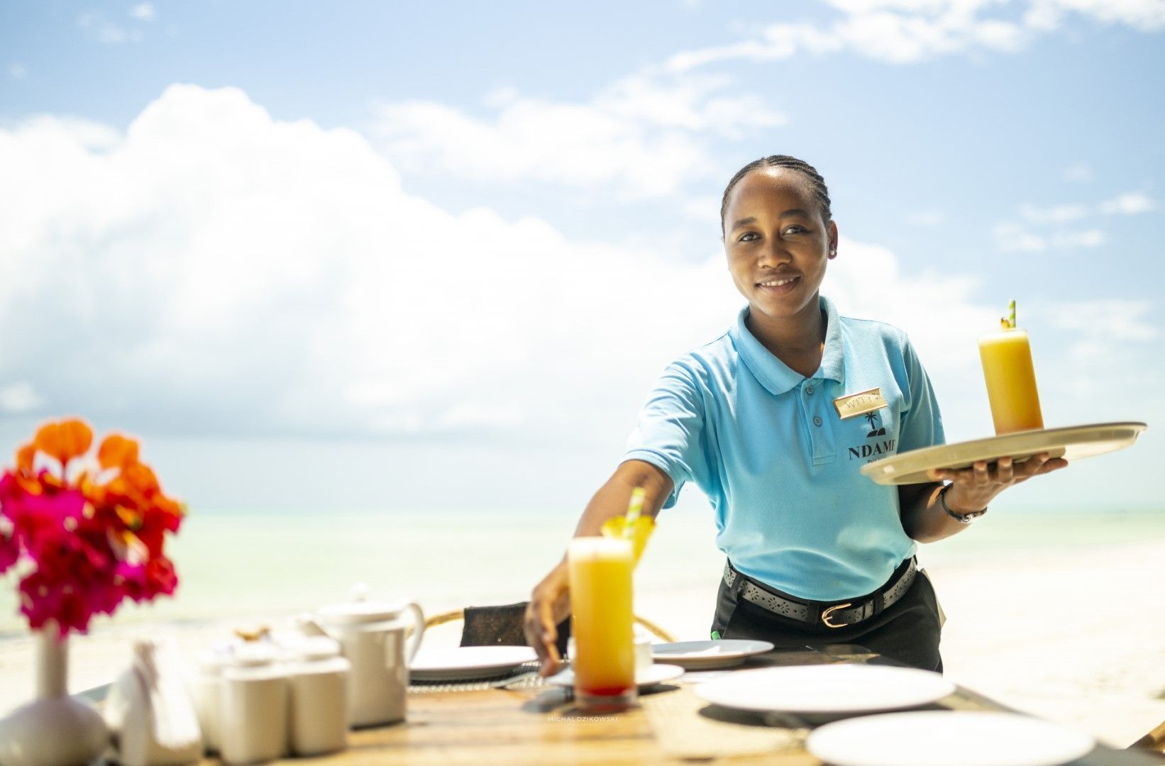 Waitress in a light blue shirt serving orange juice outdoors by the beach.