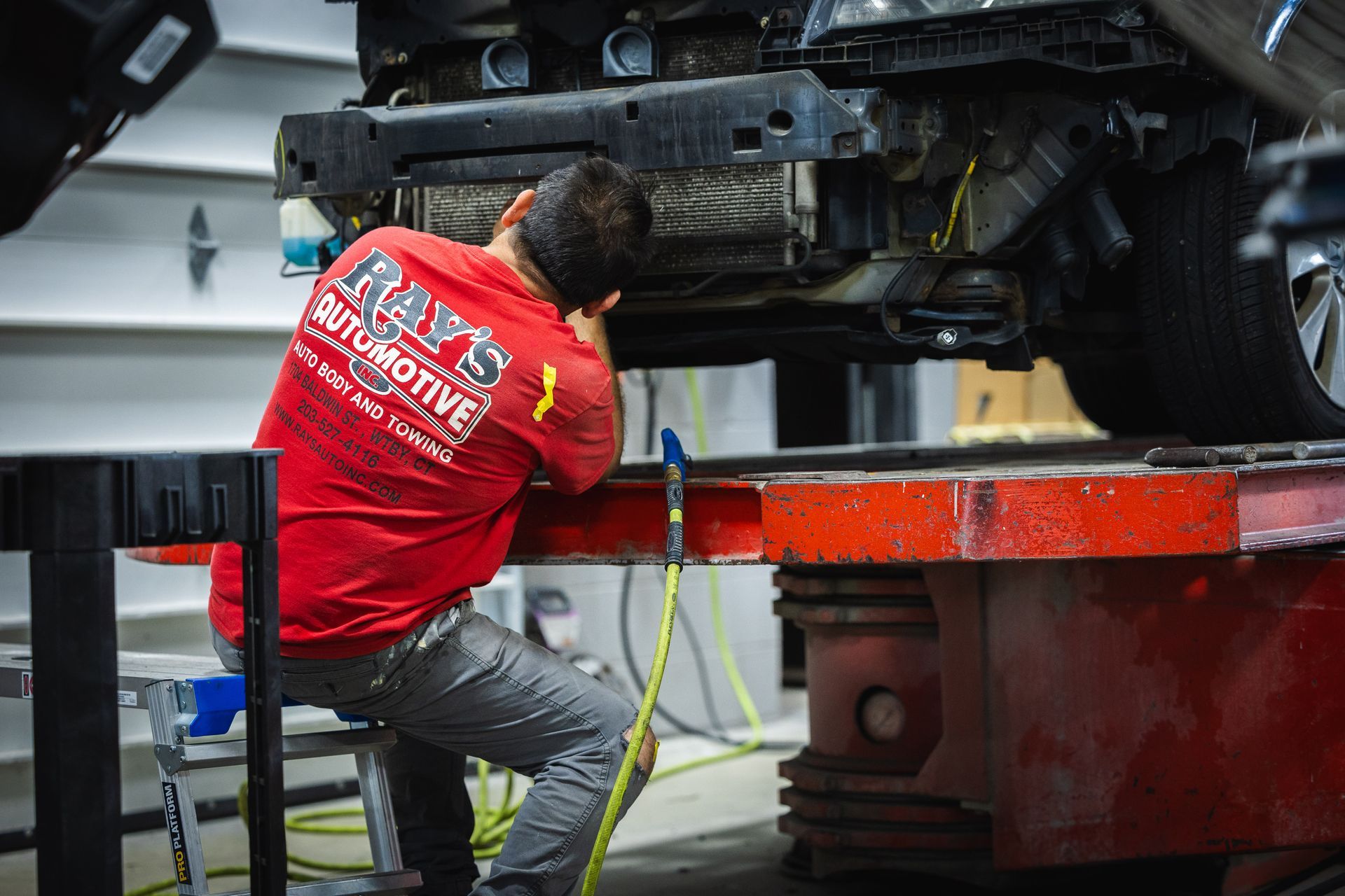 Mechanic in red shirt works under a raised car in an auto repair shop