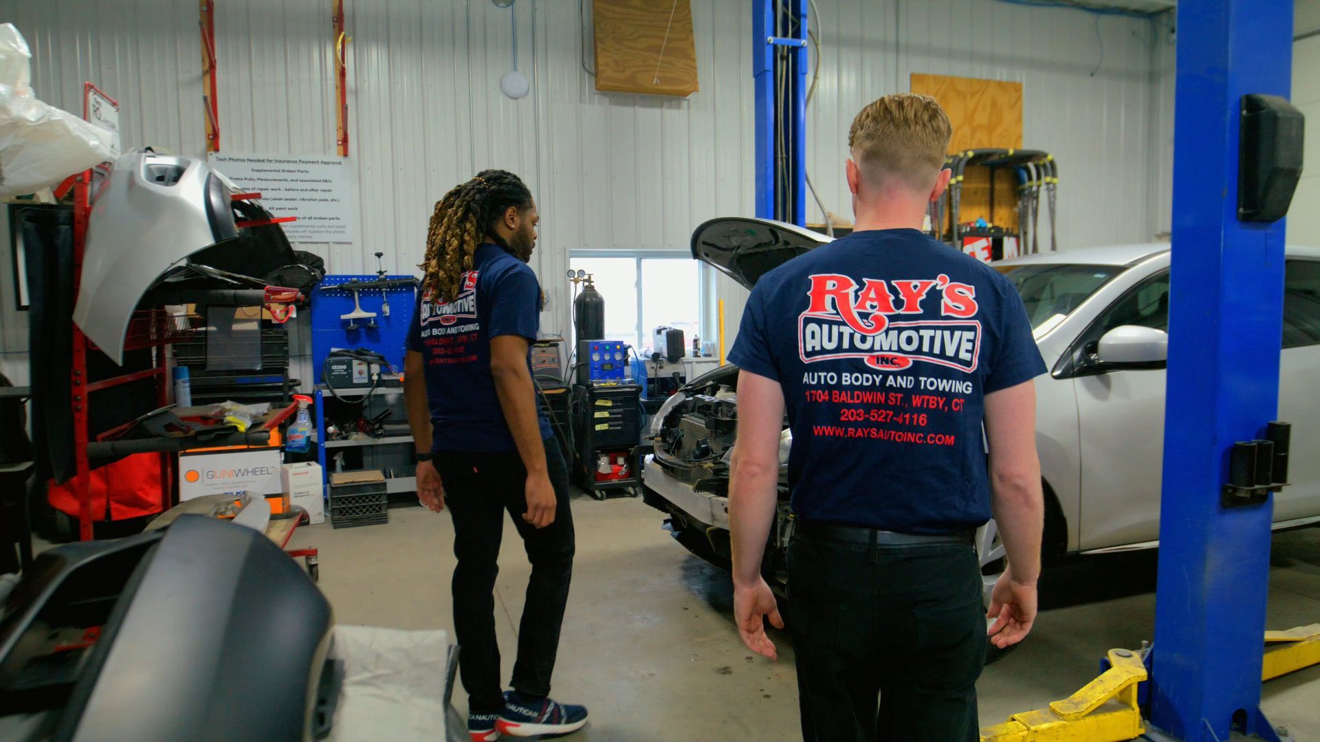 Two people in a car repair shop standing near a vehicle on a lift and tools.