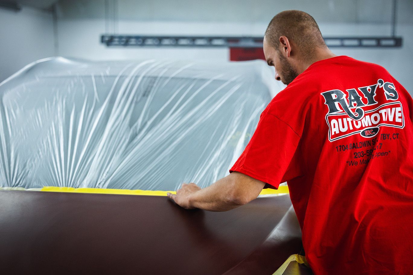 Man in red shirt applying masking tape to a car covered in plastic in an auto body shop