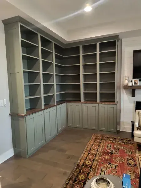 Built-in corner bookshelves with sage cabinets in a living room, beside a patterned rug and fireplace.