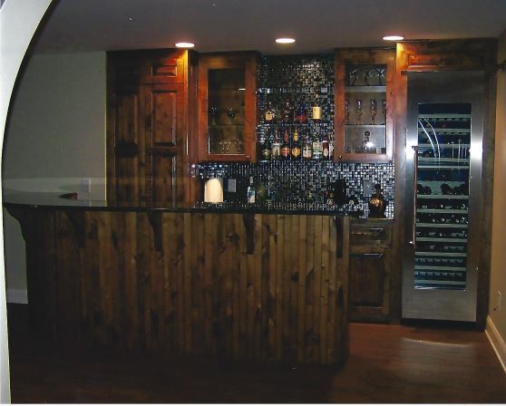 Dimly lit rustic bar with wood paneling, mosaic backsplash, and a glass-front wine cooler.