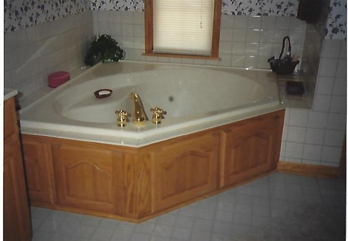 Corner bathroom whirlpool tub with gold faucets, wood-paneled base, and tiled walls