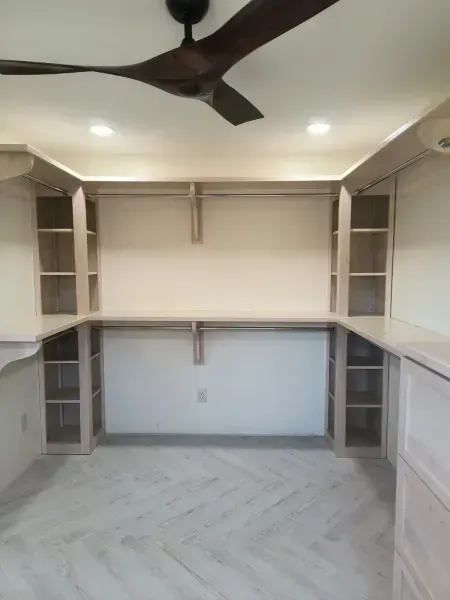 Empty walk-in closet with built-in shelves, desk, and ceiling fan on a herringbone floor.