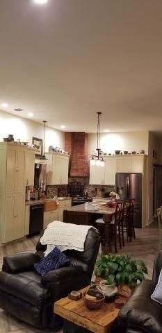 Warm open-plan kitchen and living room with a dark couch, dining table, brick chimney, and stainless fridge.