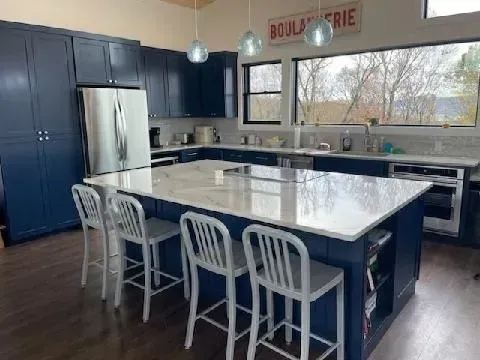 Modern kitchen with a large white island, blue cabinets, stainless fridge, and big window overlooking trees