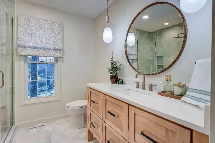 Bright bathroom with wood vanity, round mirror, hanging lights, and a window with patterned shade