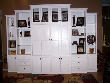 White built-in wall unit with cabinets, shelves, framed photos, and drawers in a living room