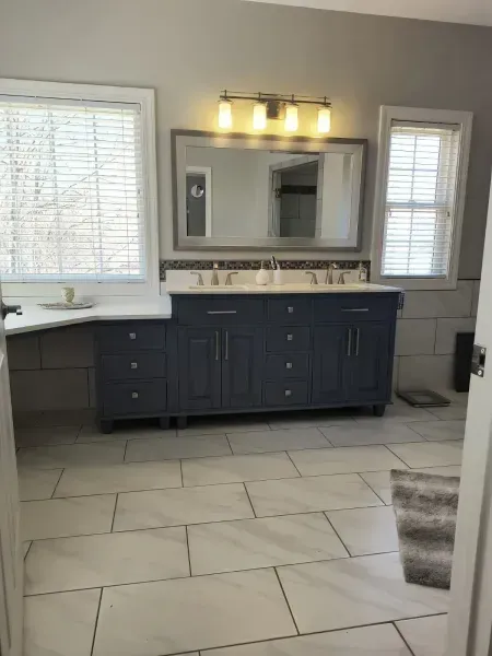 Modern bathroom with dark vanity, large mirror, white walls, tiled floor, and a window on each side of the sink.