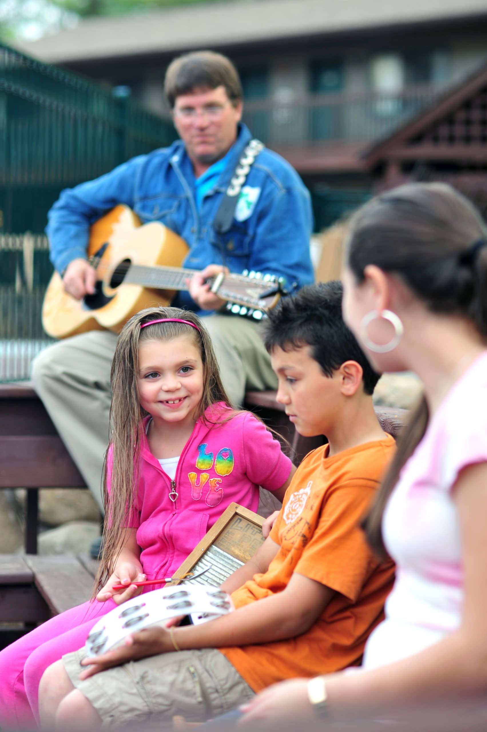 Children sit listening while a man plays guitar outdoors.