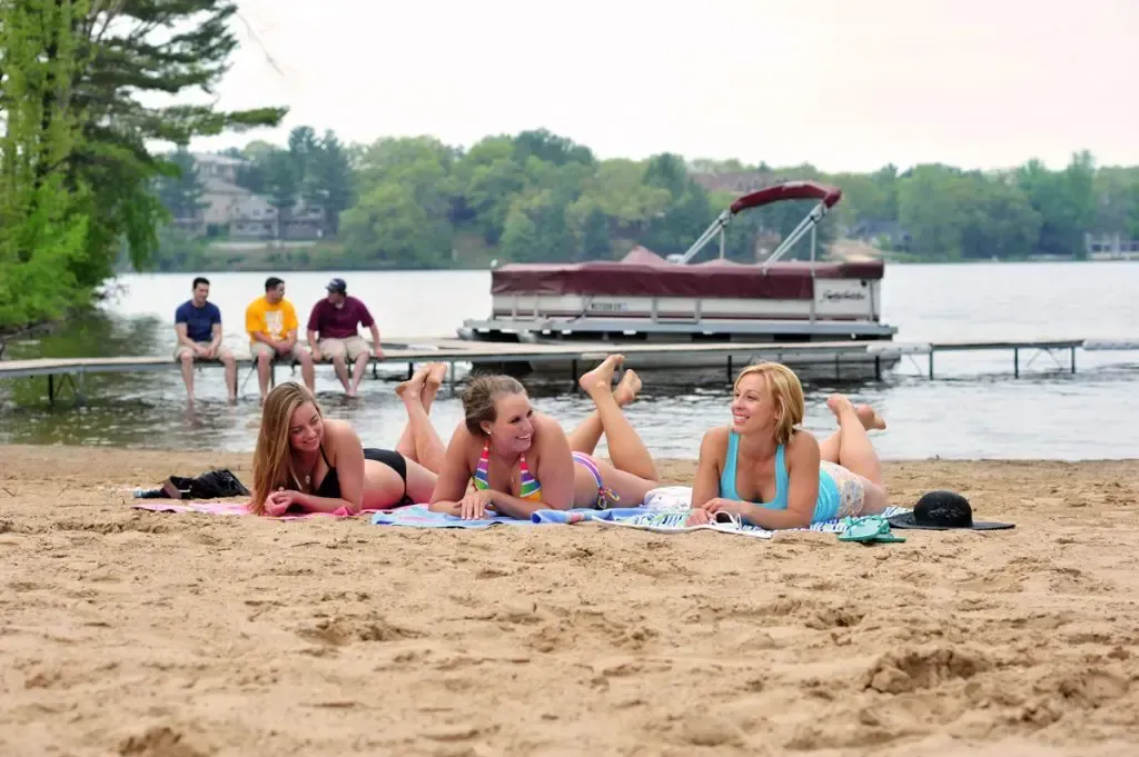 People lying on a beach towel by a lake, with a pontoon boat and dock in the background.