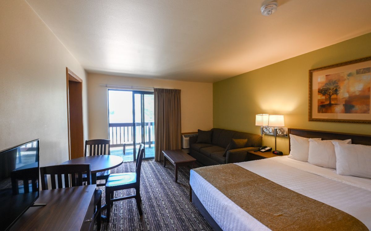 Hotel room with two beds, sofa, desk, and balcony doors, lit by warm lamps and daylight