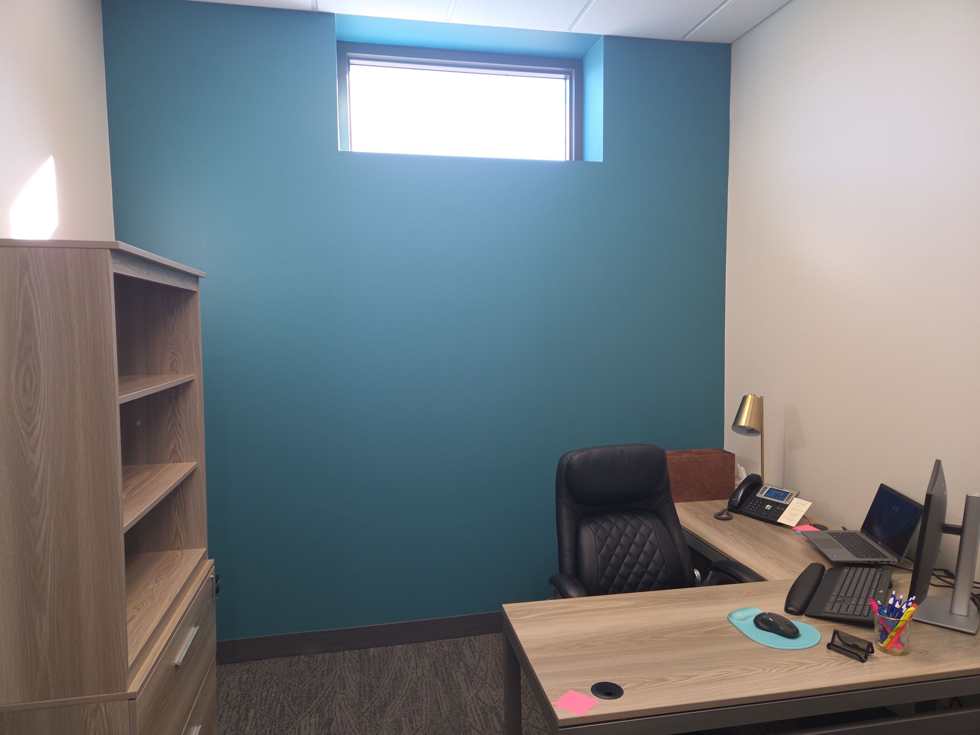 Small office with blue accent wall, desk, chair, shelving, and computer near a high window.