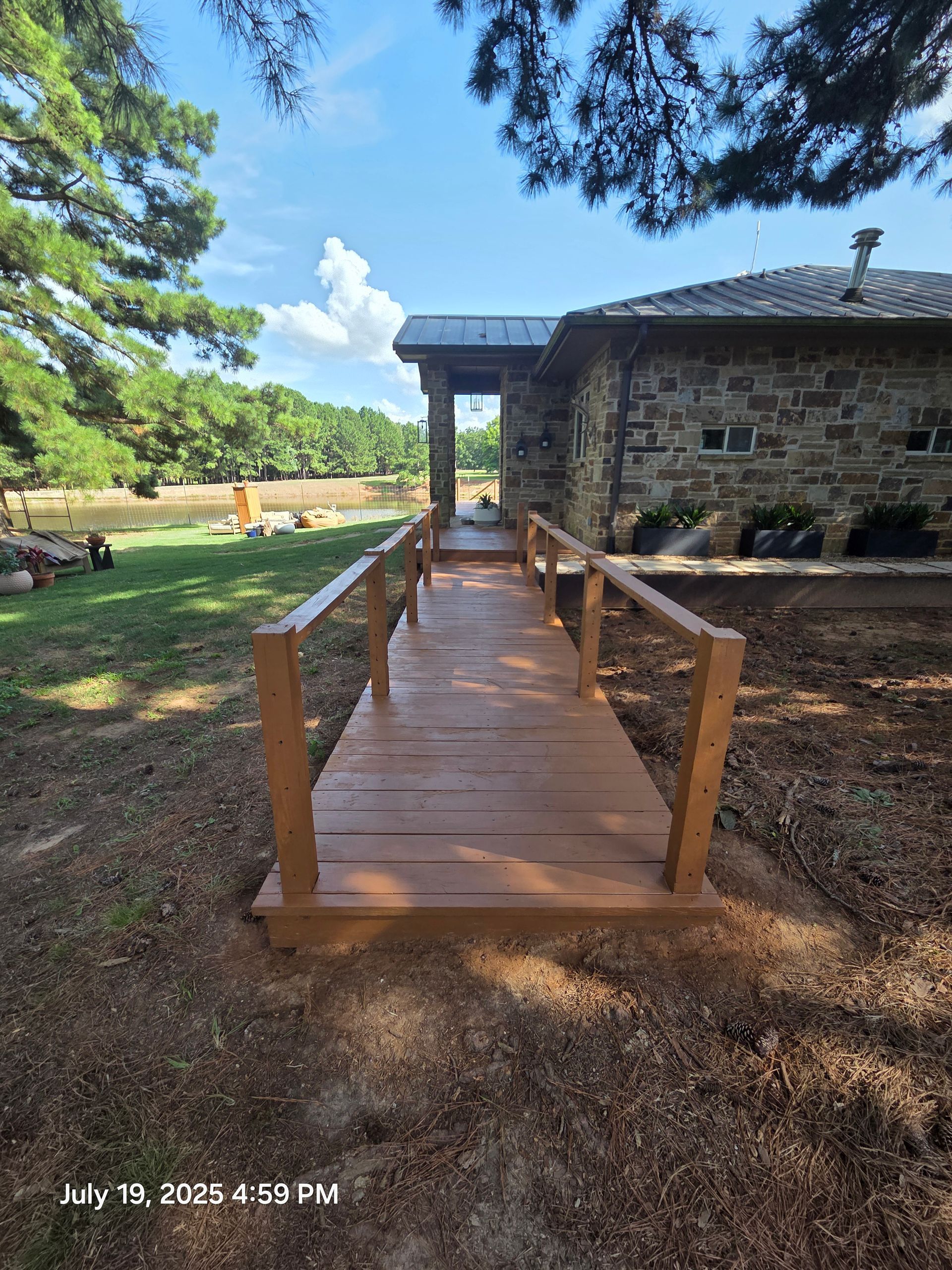 Wooden ramp leading to a stone building in a sunny park with trees and gravel path