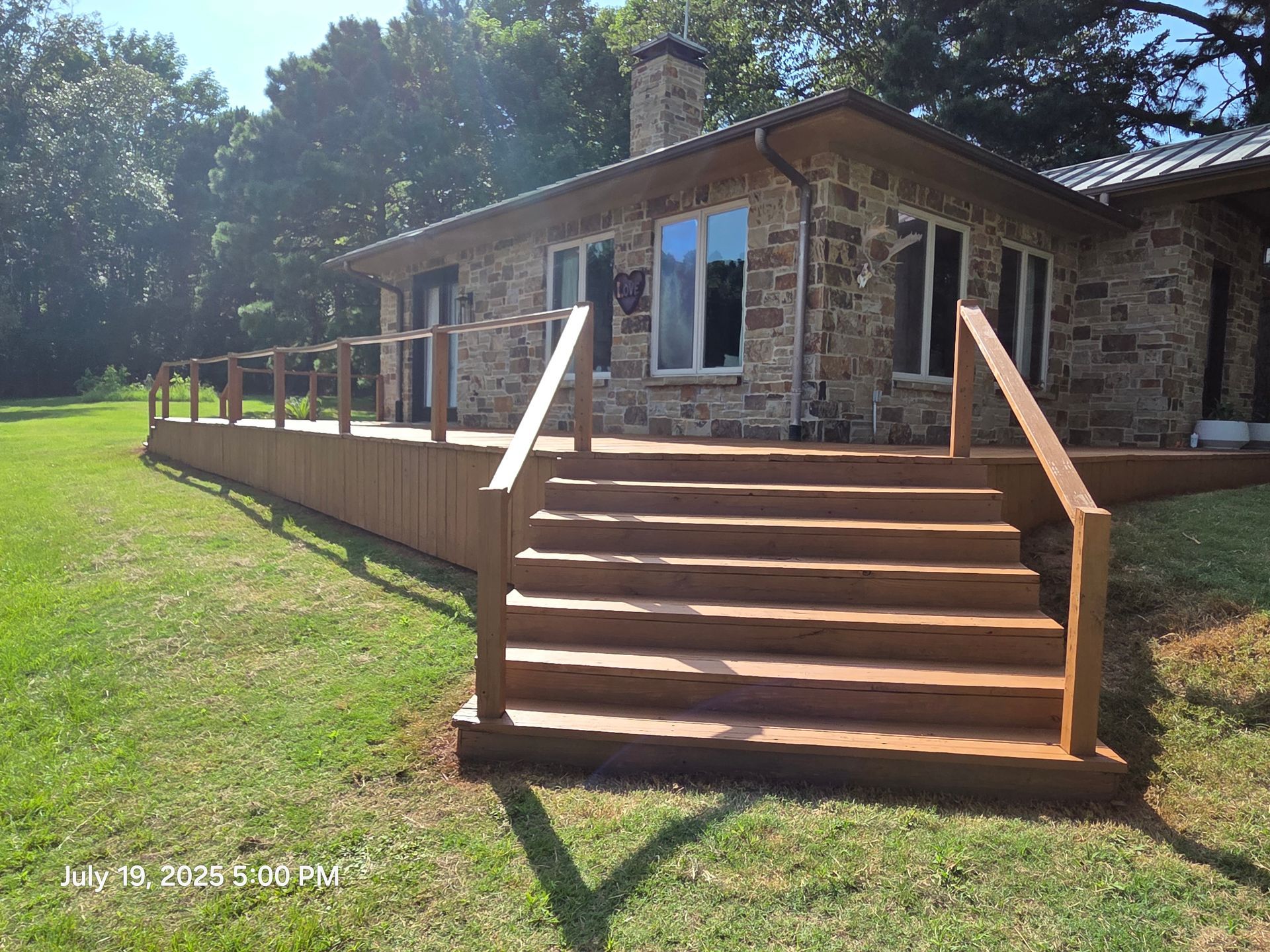 Stone house with a wooden deck, railings, and wide front steps in a grassy yard under trees.