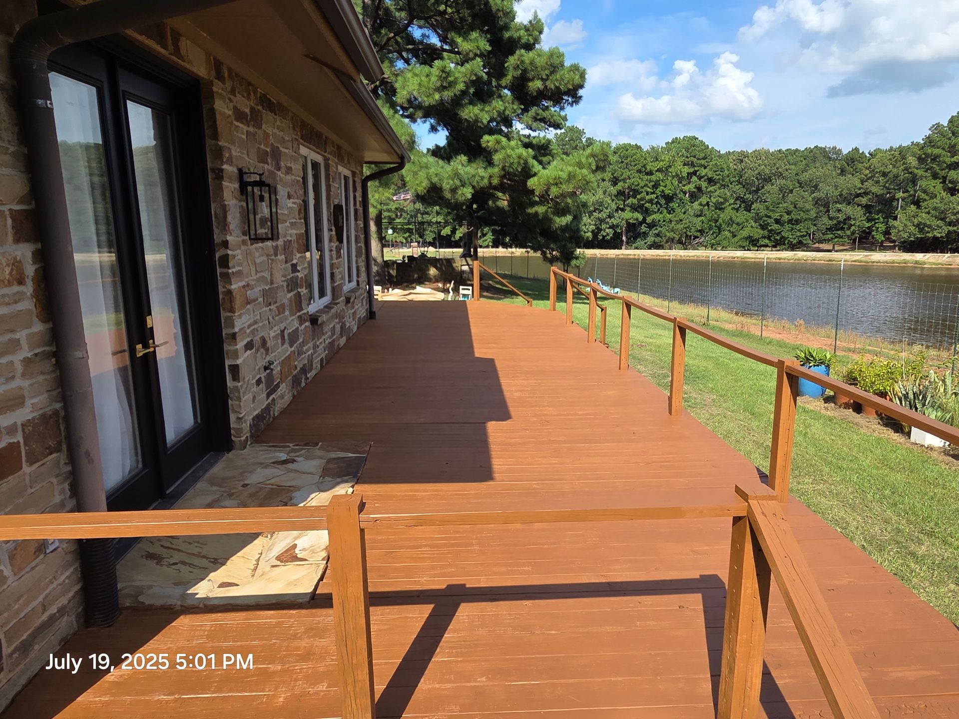 Wooden deck beside a stone house overlooking a lake, with trees and blue sky beyond.