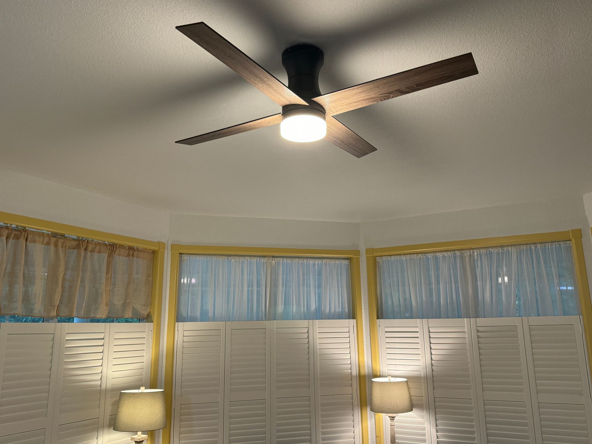 Bedroom ceiling fan with light above bay windows, white shutters, and two lamps.