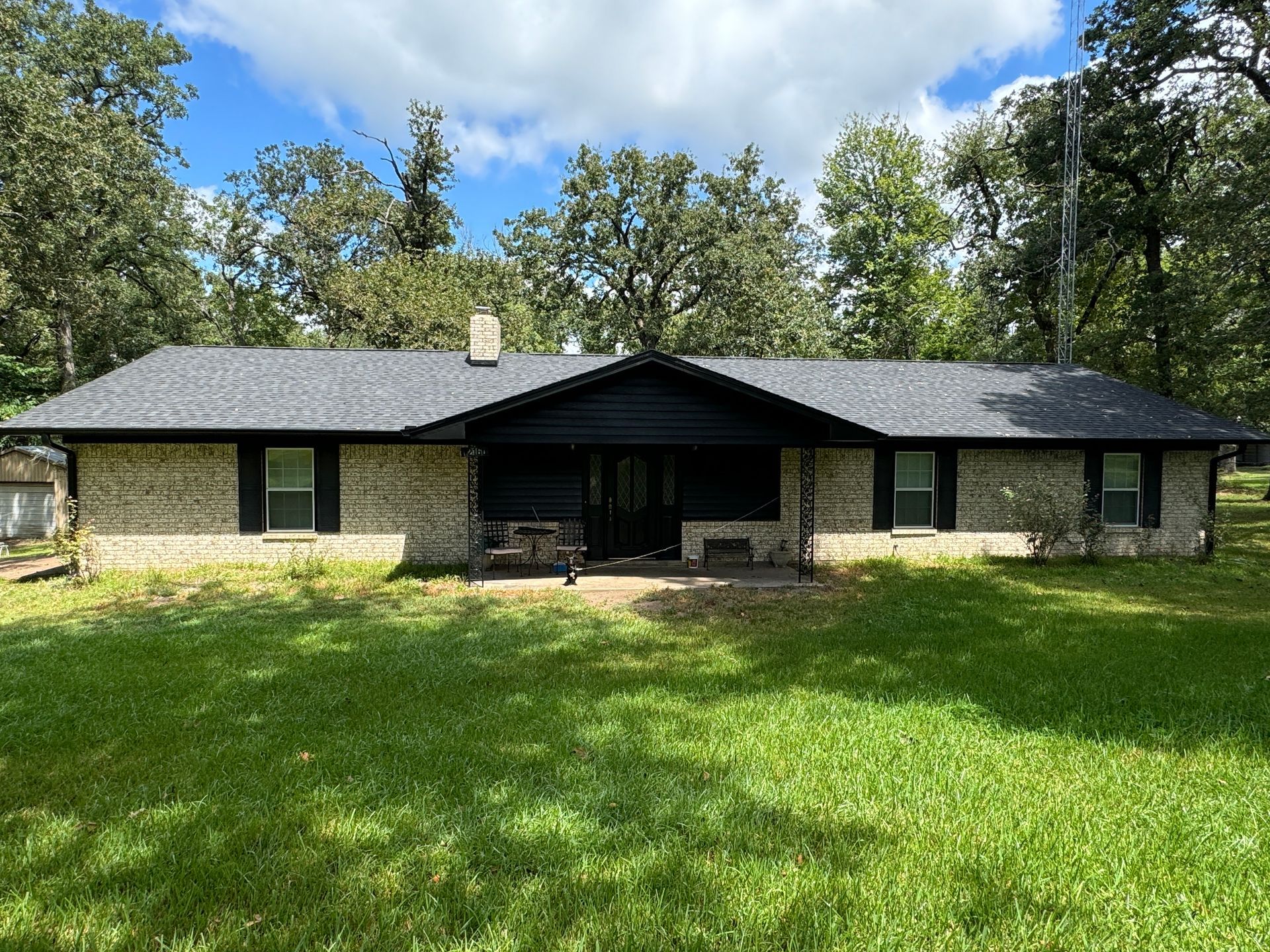 Single-story stone house with a dark roof and front porch, set in a grassy yard with trees behind it