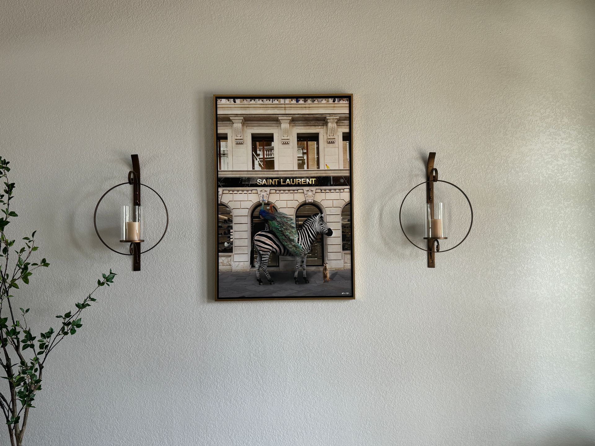 Wall art centered between two round sconces on a light textured wall, with a leafy plant at left.