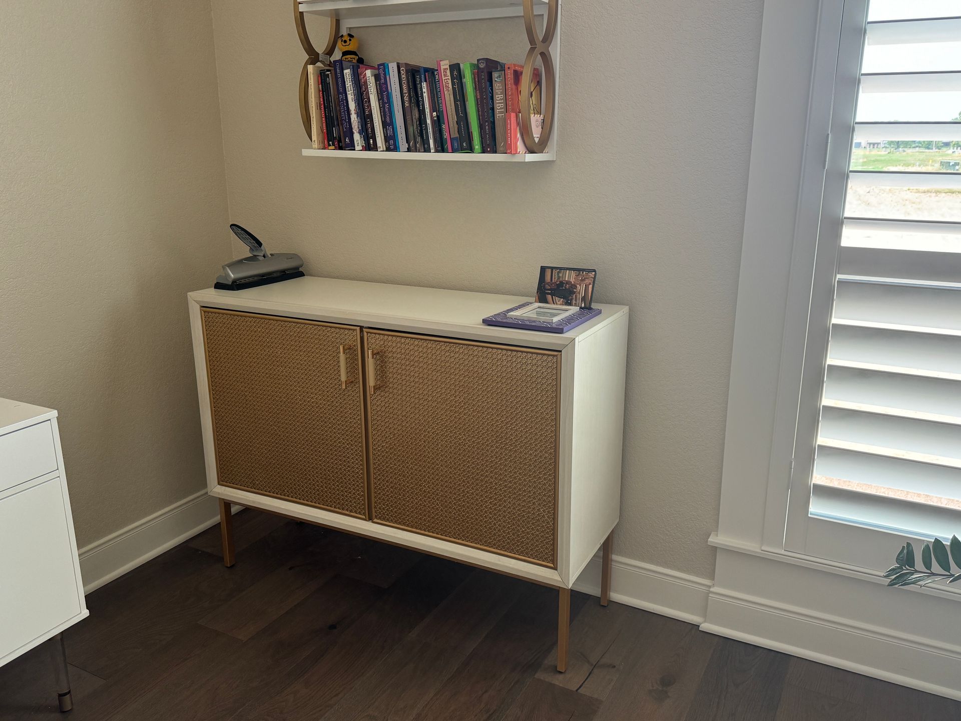 Small white cabinet with woven doors under a wall shelf in a bright room by a window.