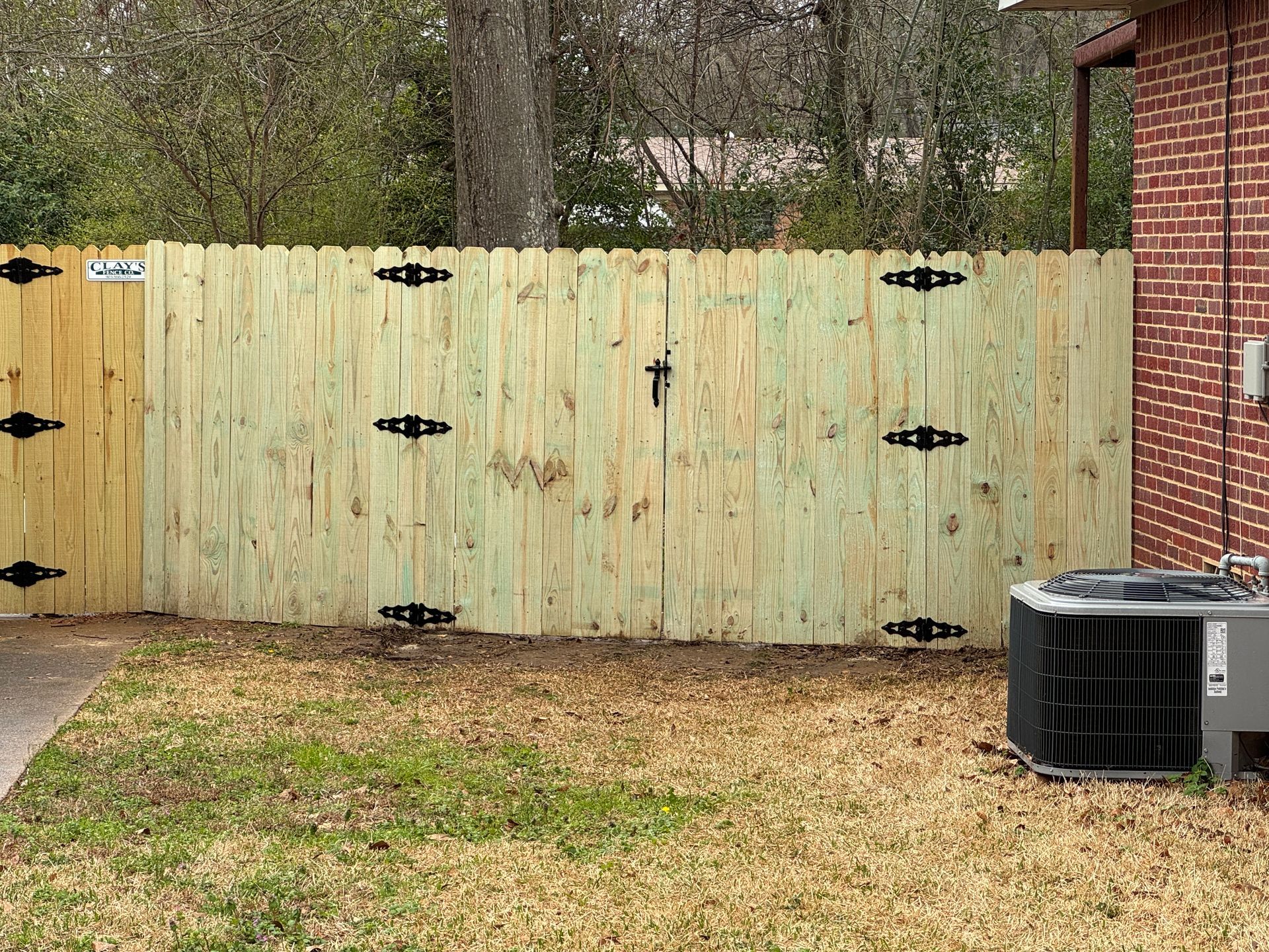 Wooden backyard gate beside a brick house, with an air conditioner unit and patchy grass in front.