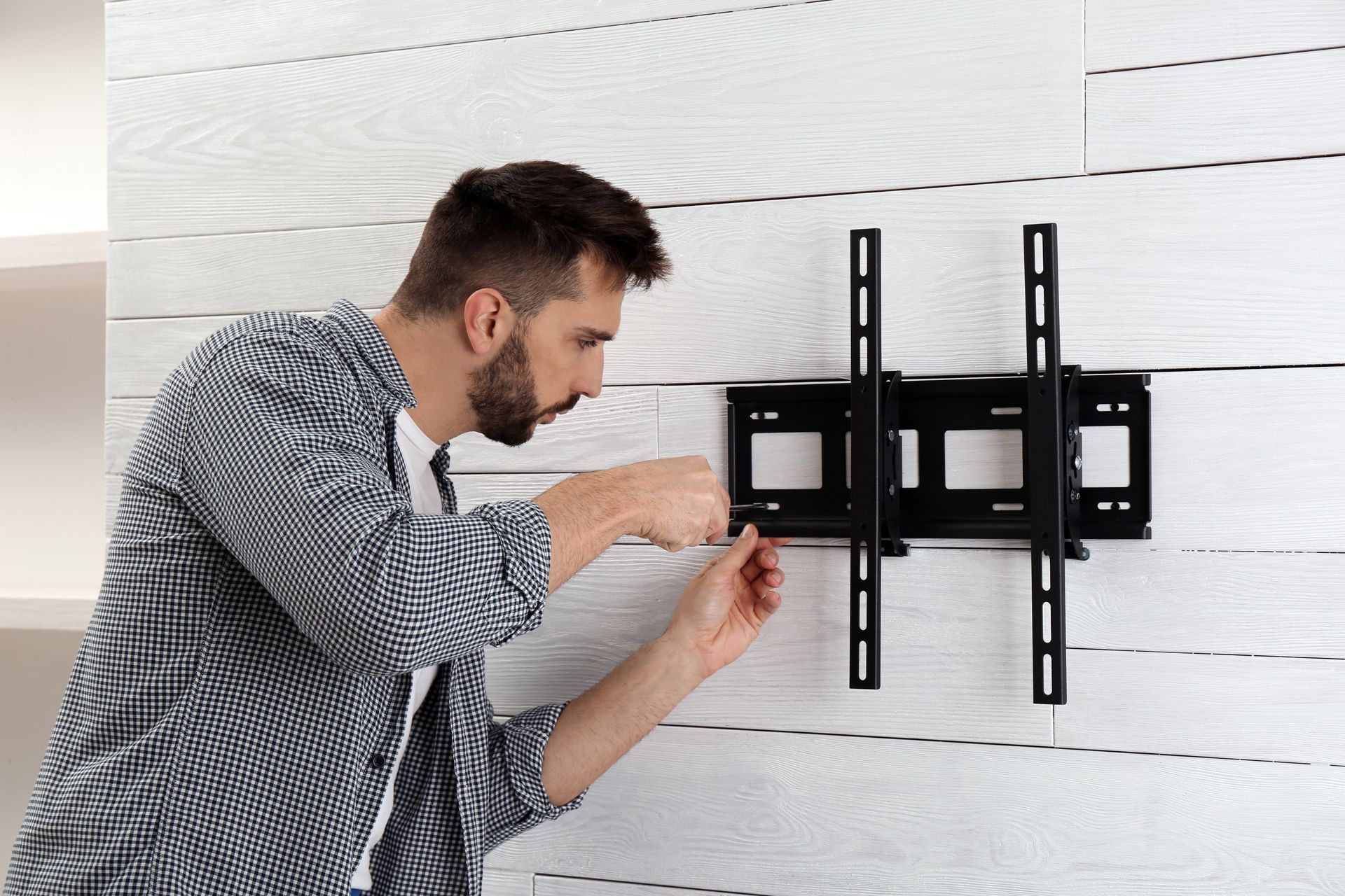Person mounting a TV bracket on a white wall, adjusting the black wall mount with a screwdriver.