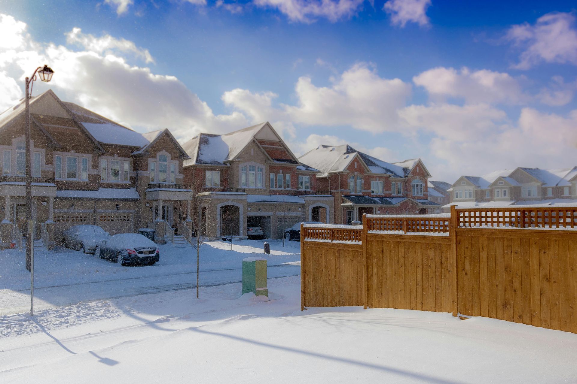 Snow-covered suburban street with row houses, parked cars, and a wooden fence under a blue sky