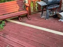 Red wooden deck with bench, grill, and railing next to green foliage.