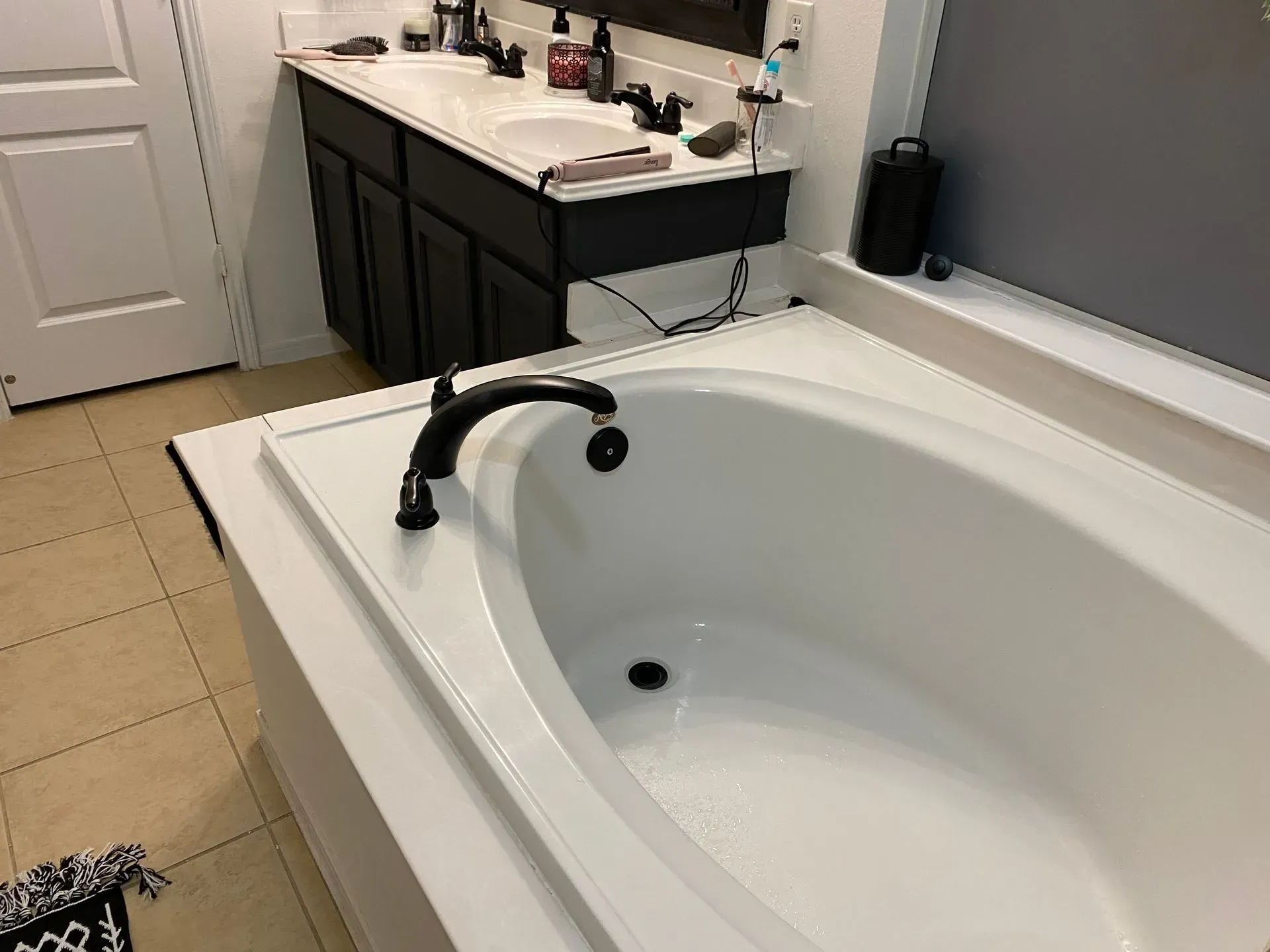 White bathtub with a black faucet beside a bathroom vanity and beige tile floor.