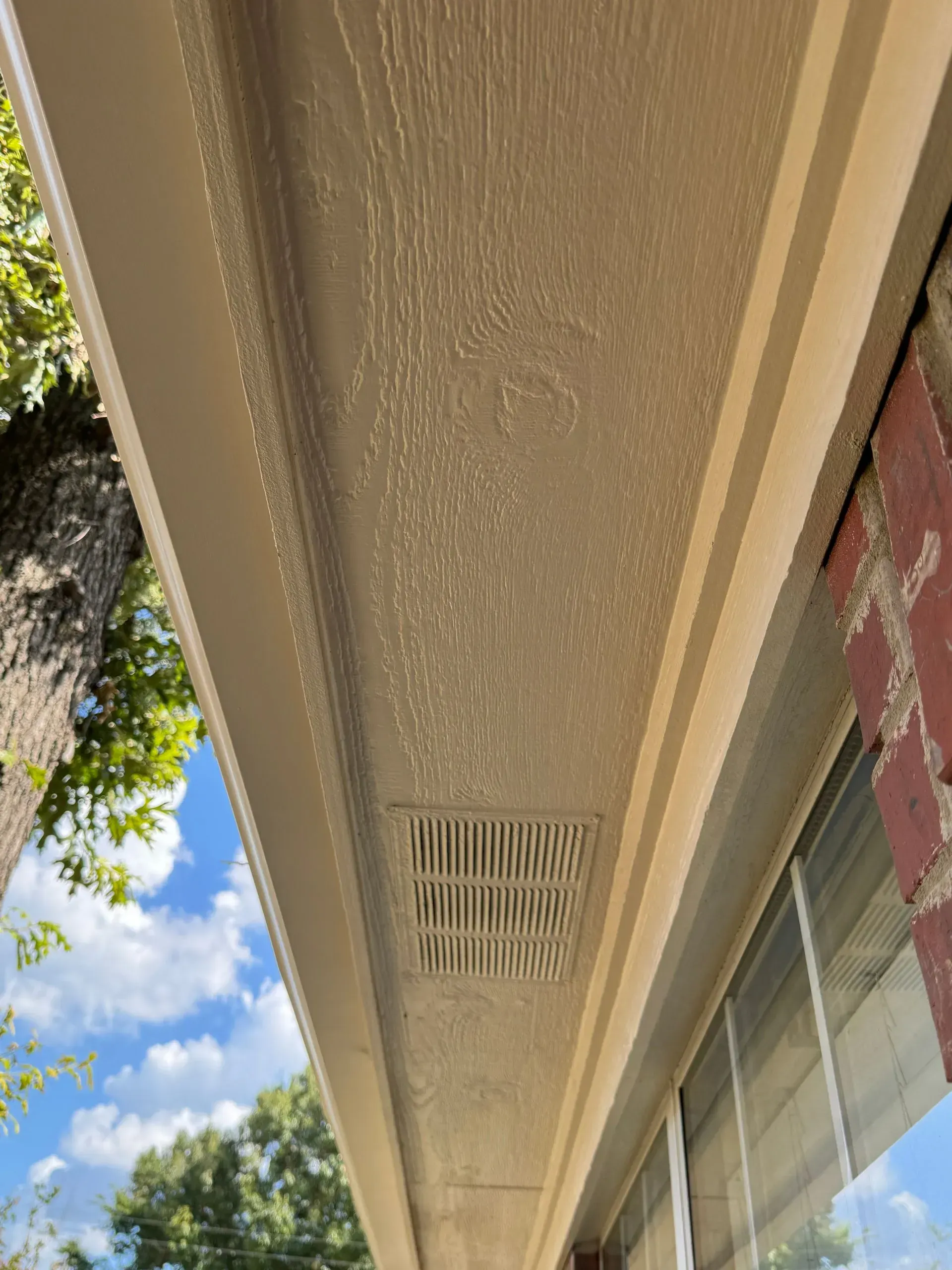 Exterior house eave with tan textured soffit, vent, and window edge beside blue sky and trees