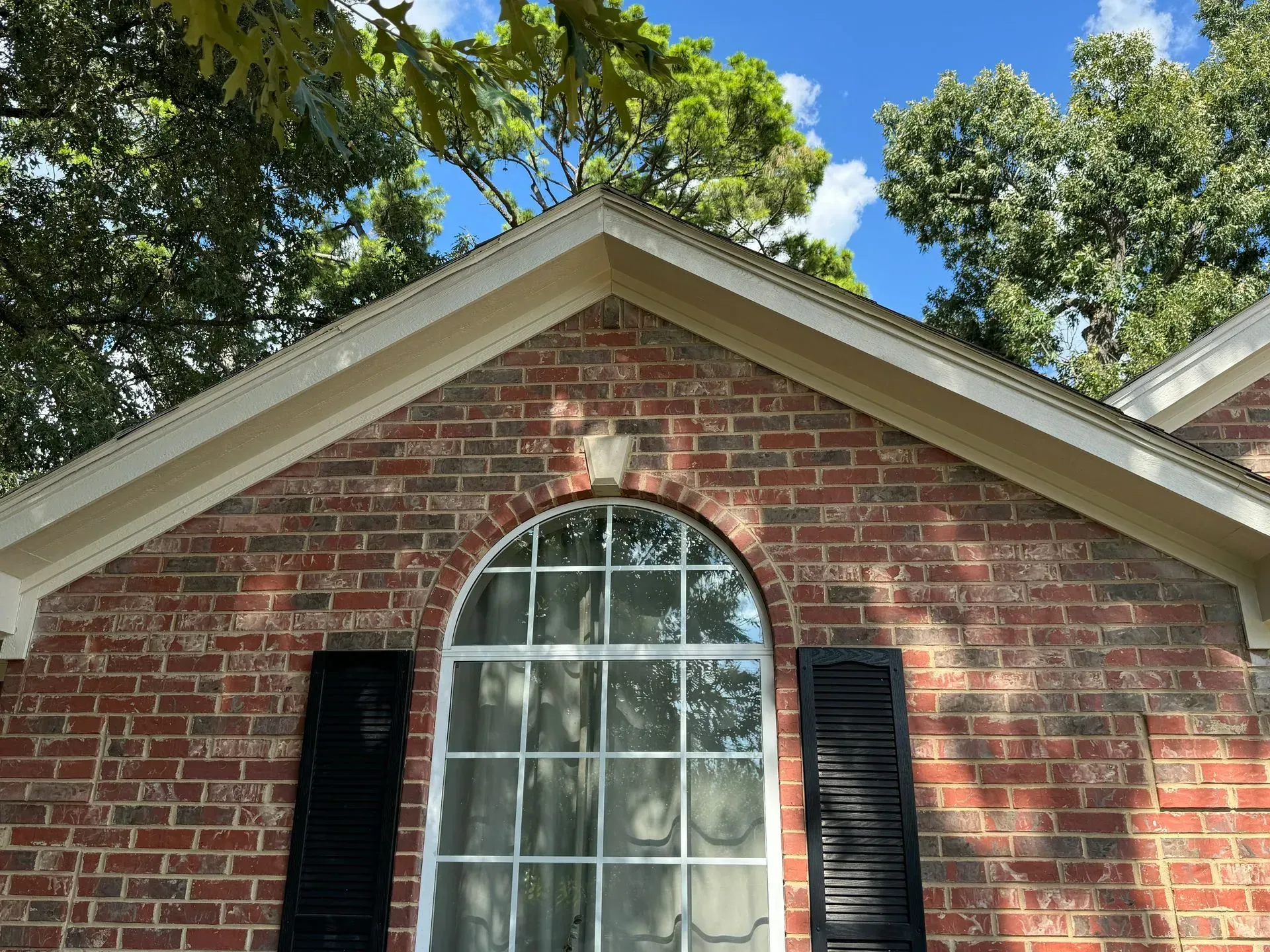 Brick house gable with arched window, black shutters, and trees overhead