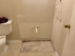 Small beige bathroom with toilet, wall plumbing, and a rough floor patch where a fixture was removed