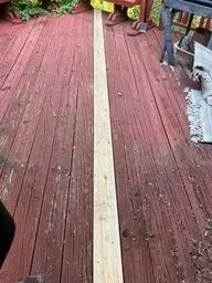 Red wooden deck with a light-colored strip running down the center, bordered by plants and gravel.