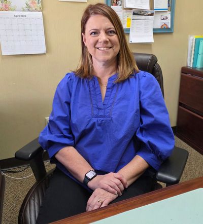 A woman is sitting at a desk with her hands folded and smiling.