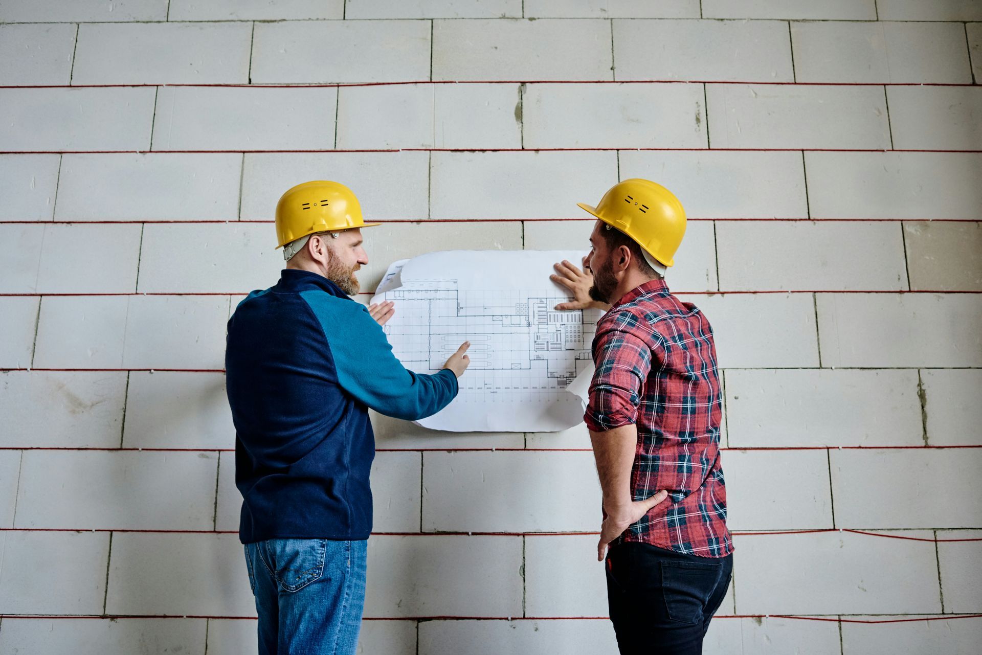Two construction workers are looking at a blueprint on a wall.