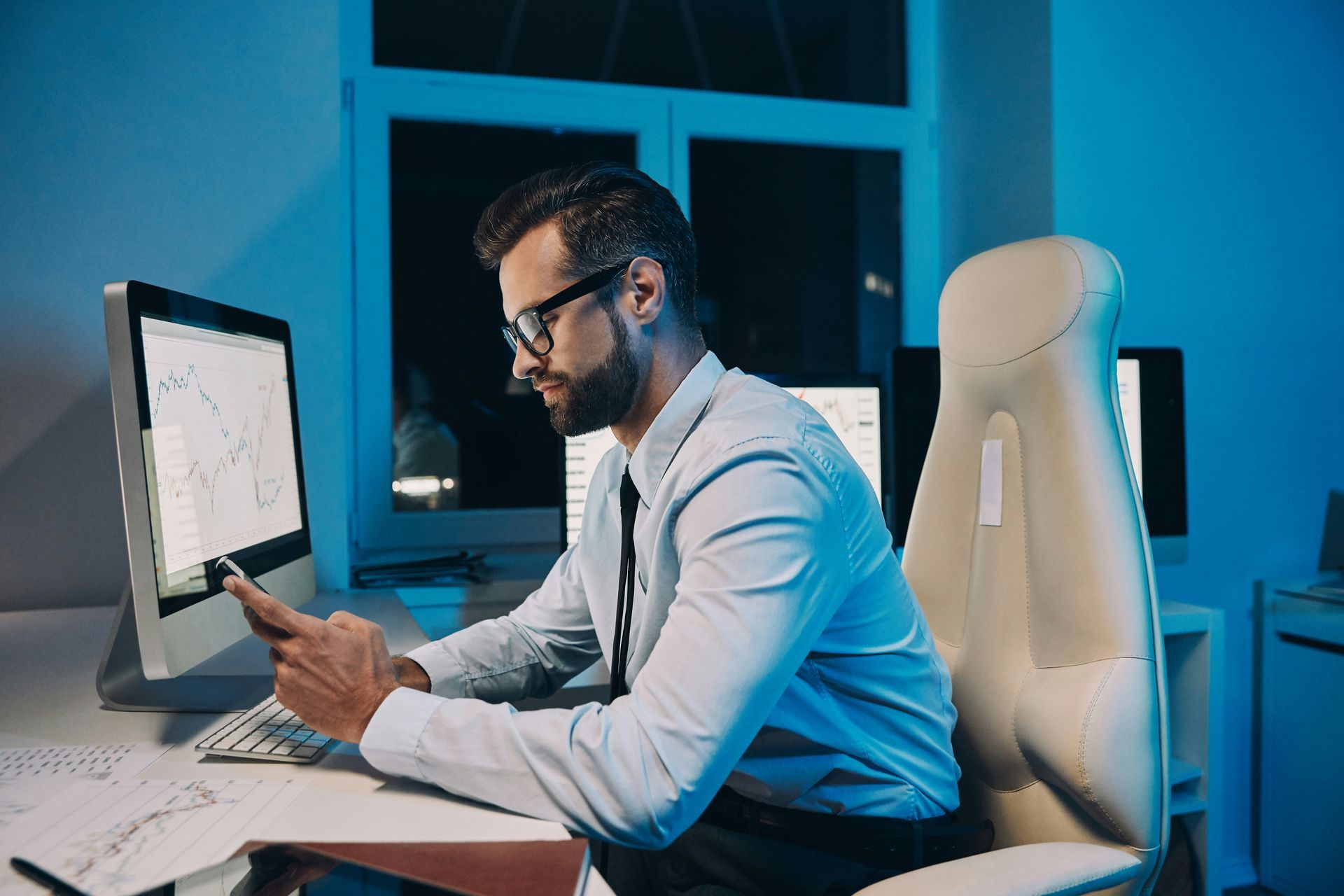 A man is sitting at a desk in front of a computer and looking at his phone.