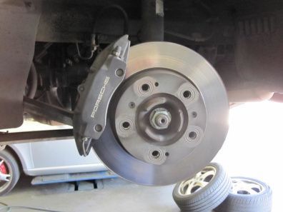 Car wheel with brake rotor and caliper visible, car raised on jack; spare tire on the ground nearby