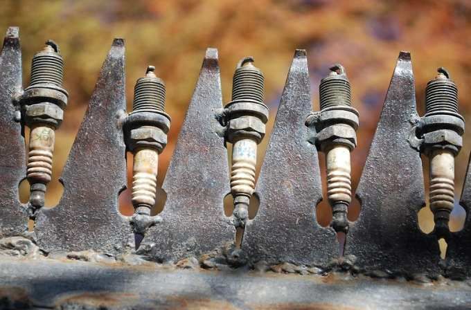 Five dirty spark plugs lined up on a dark surface, with one showing a tan ceramic insulator.